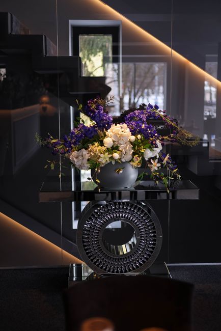 A sophisticated floral arrangement featuring white roses, purple delphiniums, and greenery, displayed in a modern, round grey vase placed on a black glossy table. The bouquet is lush and abundant, wit