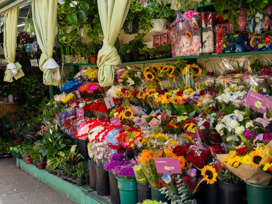 A vibrant display of floral arrangements outside a florist shop featuring a variety of fresh flowers including sunflowers, white lilies, red roses, pink carnations, purple statice, and mixed seasonal 