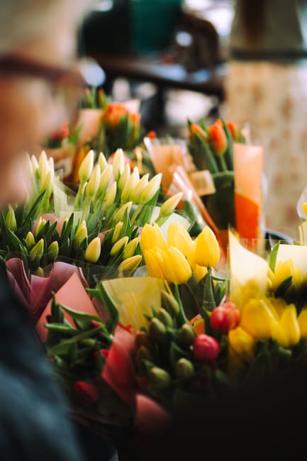 A vibrant display of fresh flowers at a florist shop, featuring bouquets with vibrant yellow tulips, white lilies, and pink buds wrapped in pastel-colored floral paper. The flowers are arranged on a c